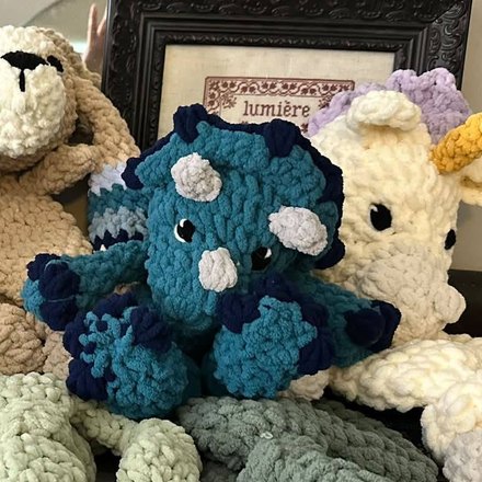 A blue crocheted triceratops lovey sitting between a bunny and a unicorn