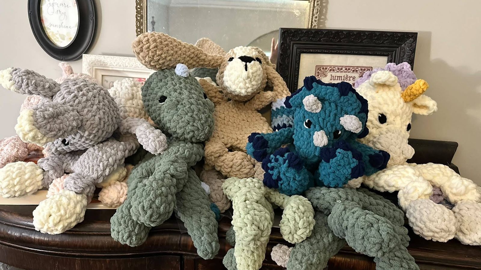 A row of handmade crochet lovey animals in chunky chenille yarn — a grey elephant, sage green dinosaur, tan bunny, teal and navy triceratops, and cream unicorn — resting on an antique wooden sideboard.
