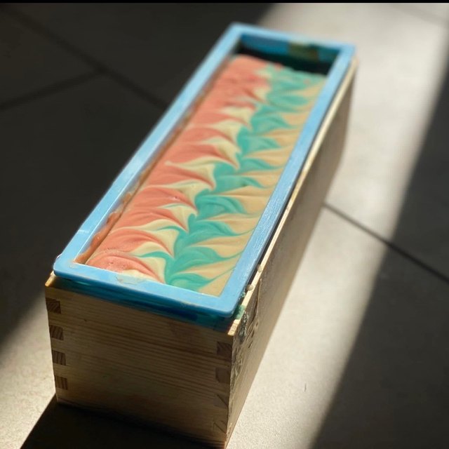 Freshly poured soap in a lined wooden mould, its surface swirled in peach, cream and teal.
