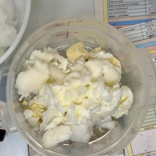 Solid white and cream oils weighed into a measuring jug, next to a printed soap recipe sheet.